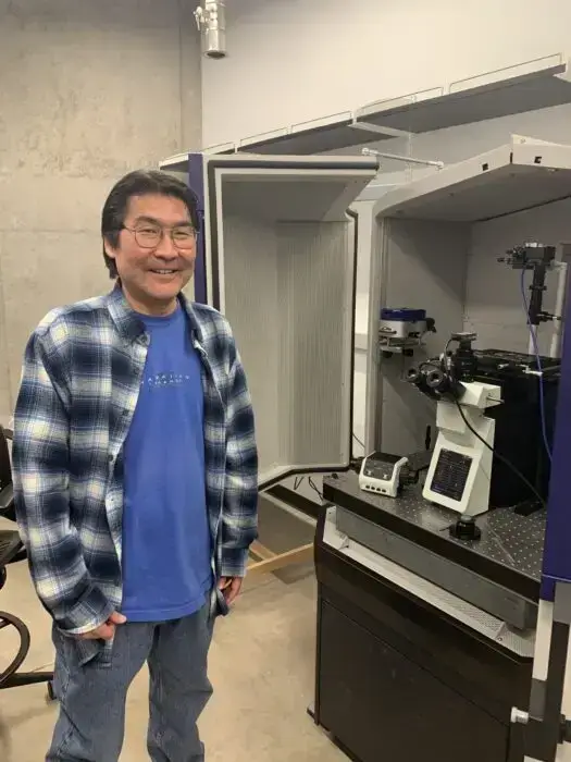 headshot of Gregory Uyeda standing next to microscope