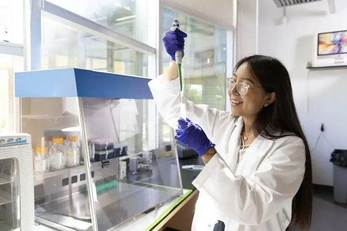 A female scientist with long black hair and glasses and gloves fills a pipette with liquid in a well-lit science lab